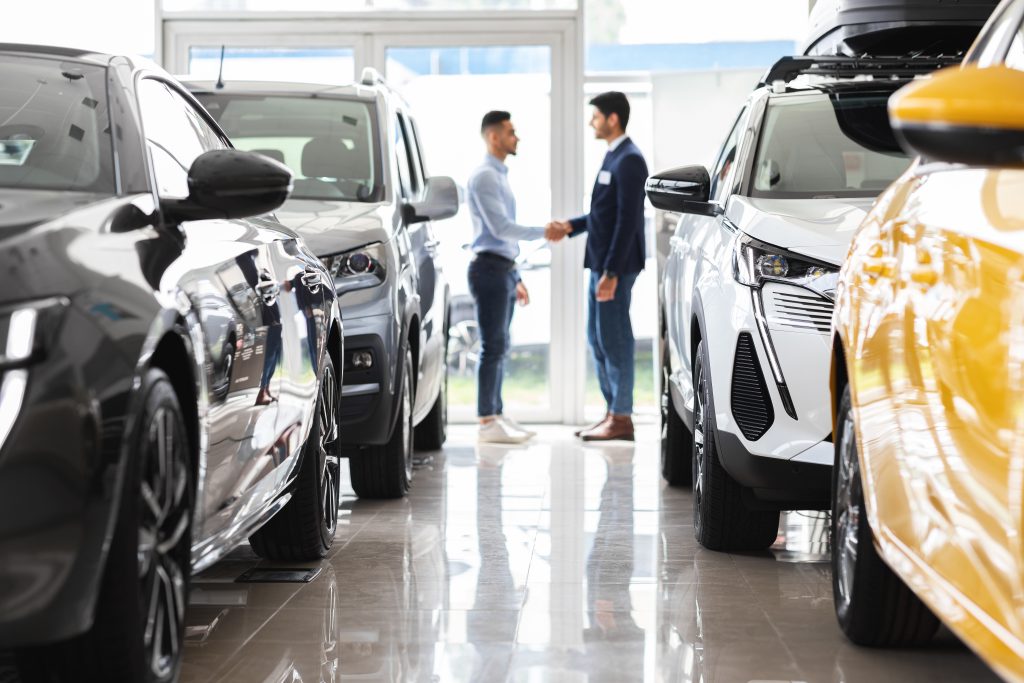 Middle-eastern young man customer and sales assistant shaking hands, car showroom interior, friendly manager greeting wealthy arab guy, standing among luxury brand new autos, full size shot